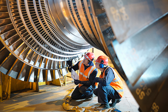 Two technicians dressed in safety gear with noise cancelling earmuffs on kneeling while they work on a turbine